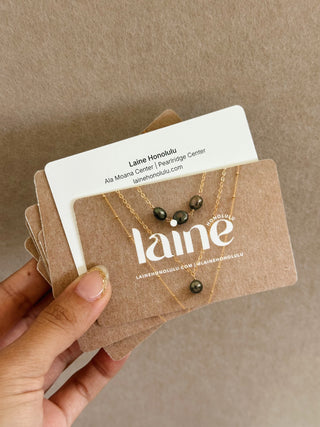 Hand holding a necklace on a 'laine' branded gift card against a brown background